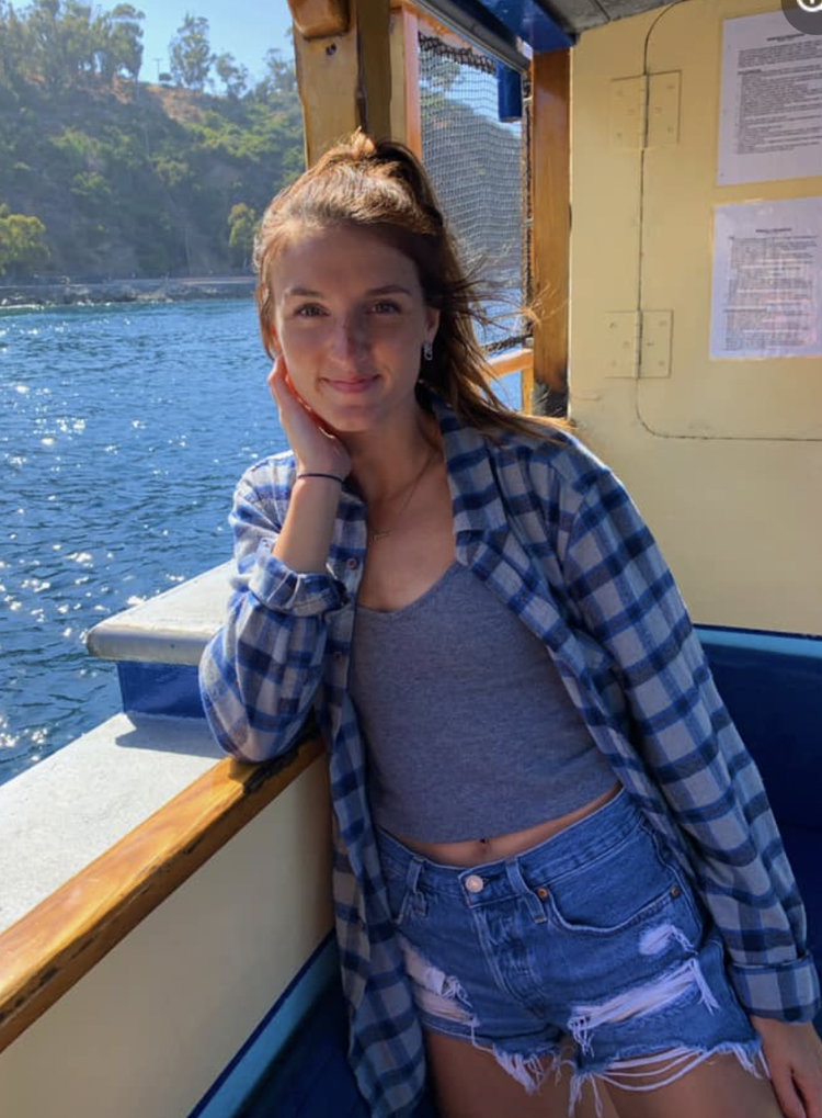 A young woman standing on a boat, leaning on the rail with her hand resting on her cheek, smiling at the camera. She has long hair pulled back, and is wearing a gray tank top, distressed denim shorts, and a blue plaid shirt. The boat is on a body of 