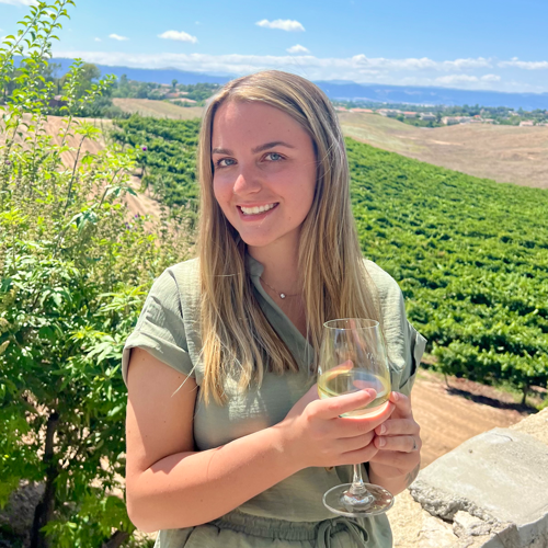 A young woman with long blonde hair smiling and holding a glass of white wine outdoors on a sunny day, with a vineyard and rolling hills in the background.