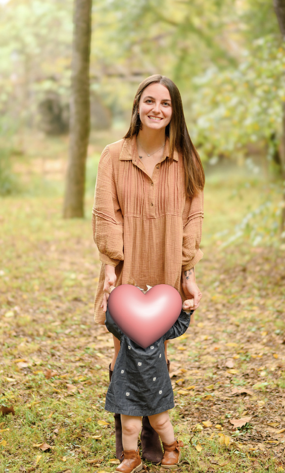 Young woman with long brown hair, wearing a light brown, long-sleeved dress, standing outside in a wooded area, holding a heart-shaped object in front of a child.