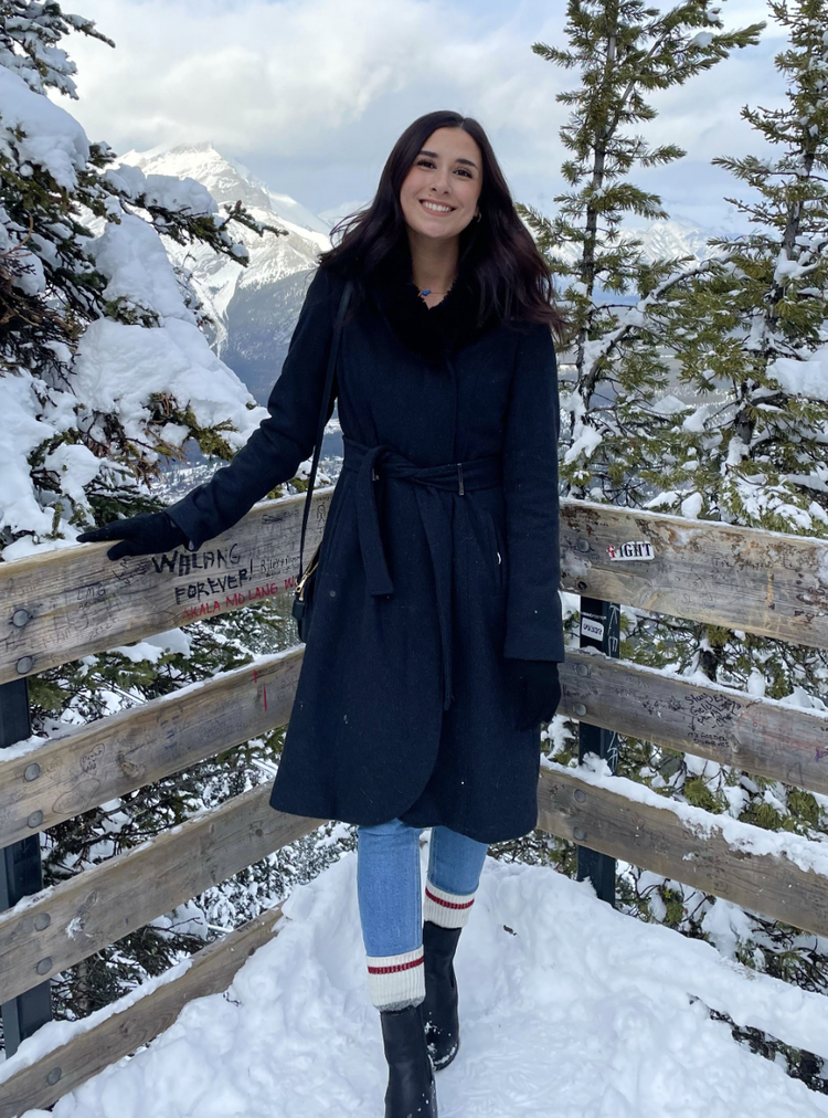 A woman with long dark hair smiling at the camera, wearing a black coat, jeans, and black boots, standing on a snowy mountain overlook surrounded by snow-covered trees and mountains in the background.