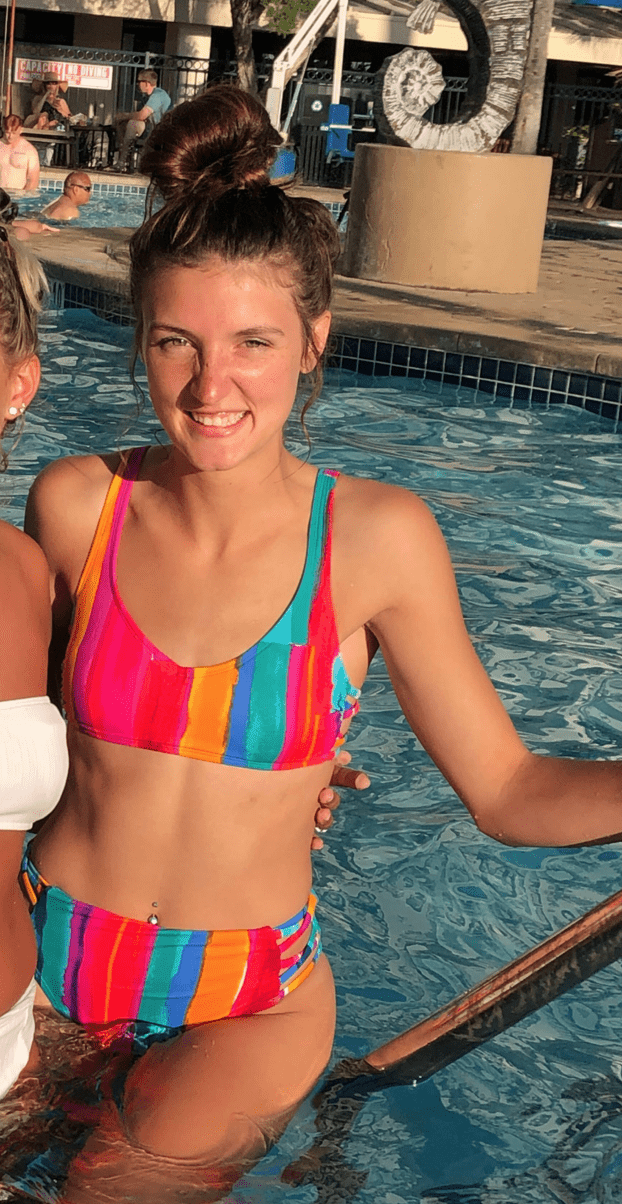 Young woman with hair in a bun, wearing a colorful rainbow striped bikini, standing in a swimming pool and smiling at the camera.