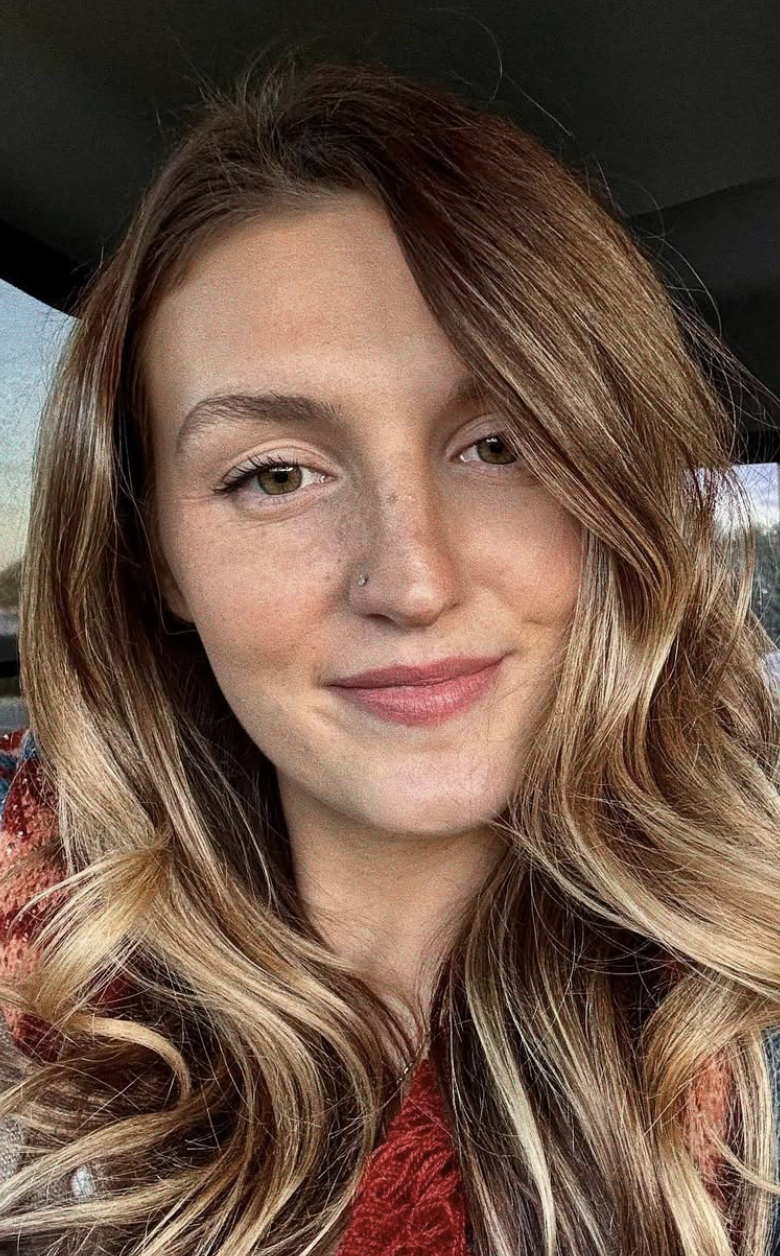 Close-up of a young woman with long wavy hair, light skin, and freckles, smiling softly while sitting in a car.