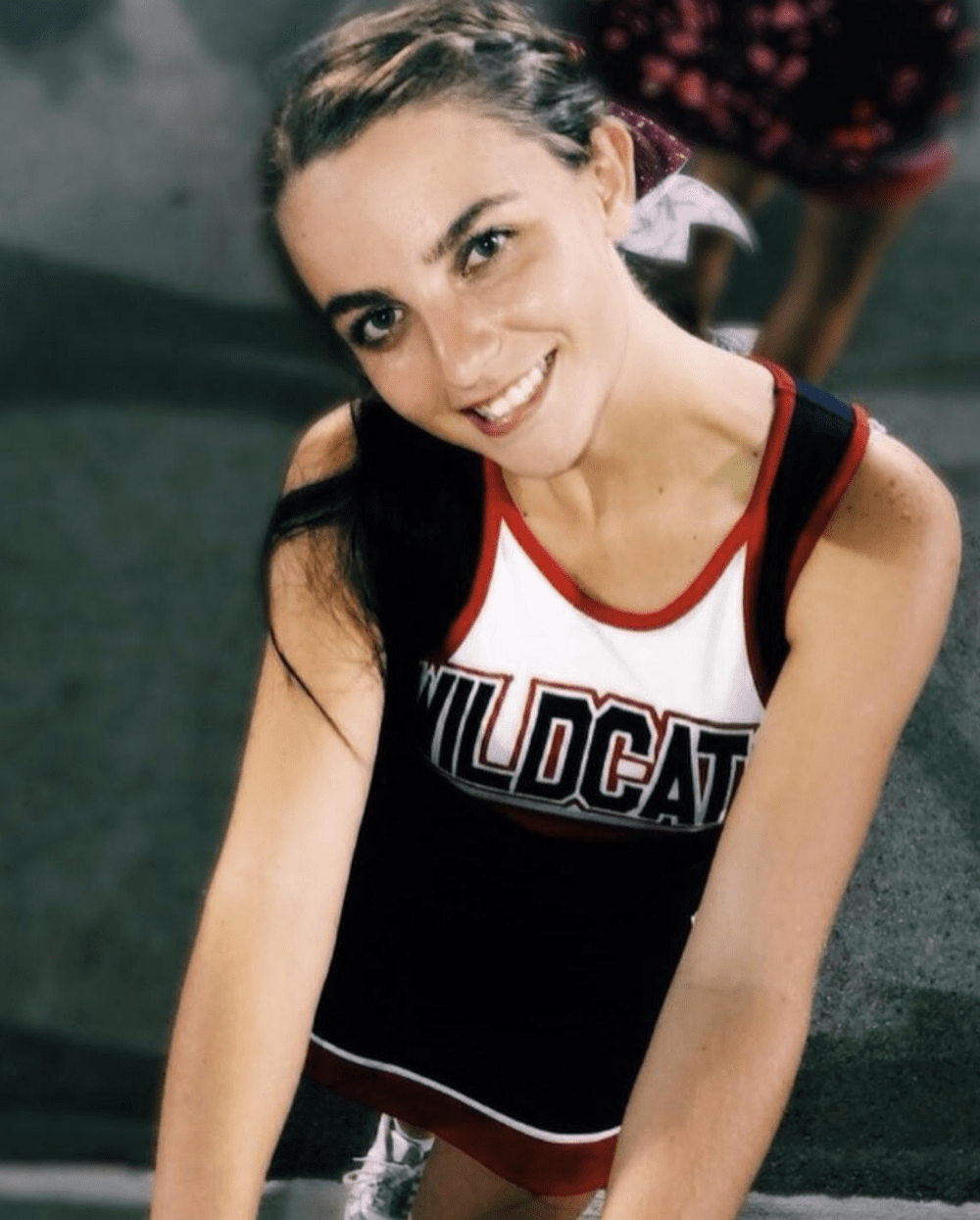 A smiling young woman in a cheerleading uniform with the word 'Wildcat' on the front, sitting on the ground with one arm extended upward.