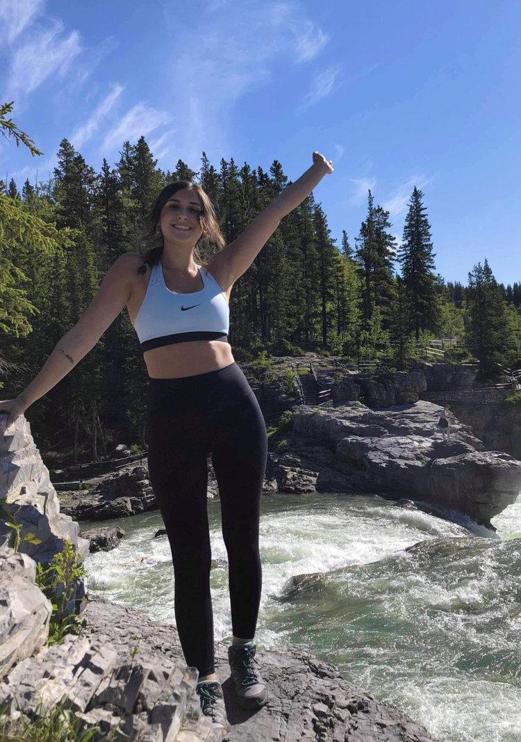 A woman in athletic wear stands on a rocky riverbank with her arm raised, smiling, surrounded by pine trees under a bright blue sky.