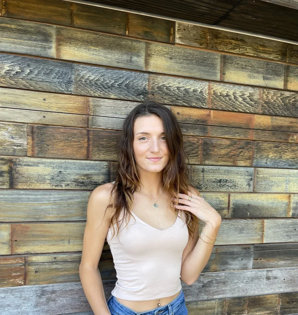 A young woman with long wavy brown hair standing in front of a rustic wooden wall, wearing a light-colored tank top and blue jeans.