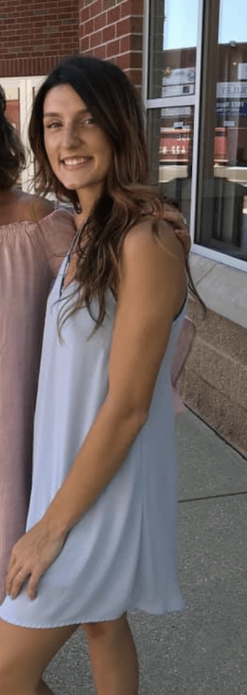 A young woman with long, wavy brown hair wearing a white dress, standing outside near a brick building with windows.