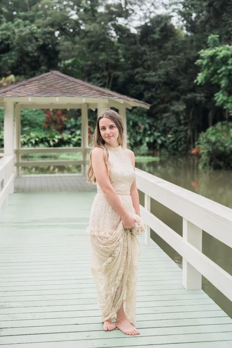 Young woman in a cream-colored lace dress standing barefoot on a wooden dock by a river, holding her dress, with a white gazebo and lush green trees in the background.