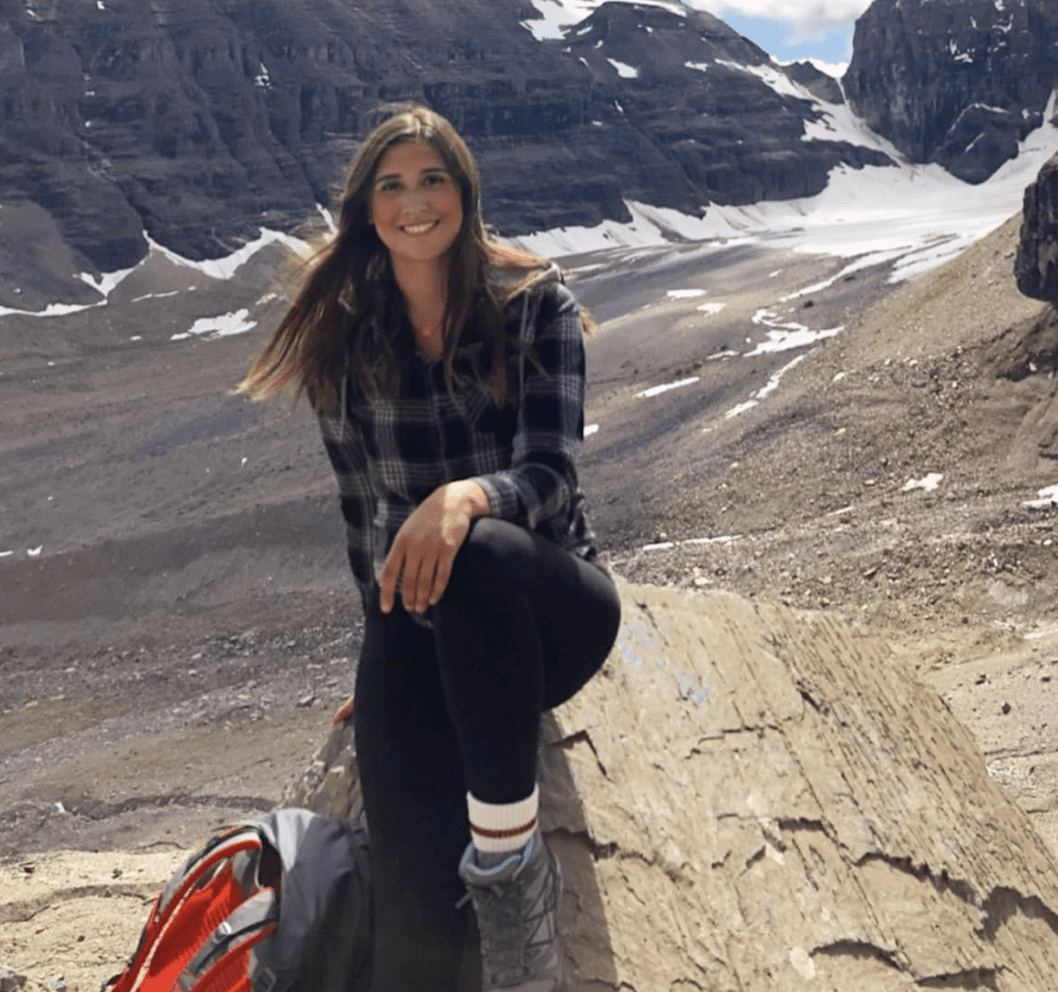 A young woman with long brown hair smiling, wearing a black plaid shirt, black pants, and gray hiking shoes, sitting on a large rock in a mountainous landscape with snow patches and rocky terrain.