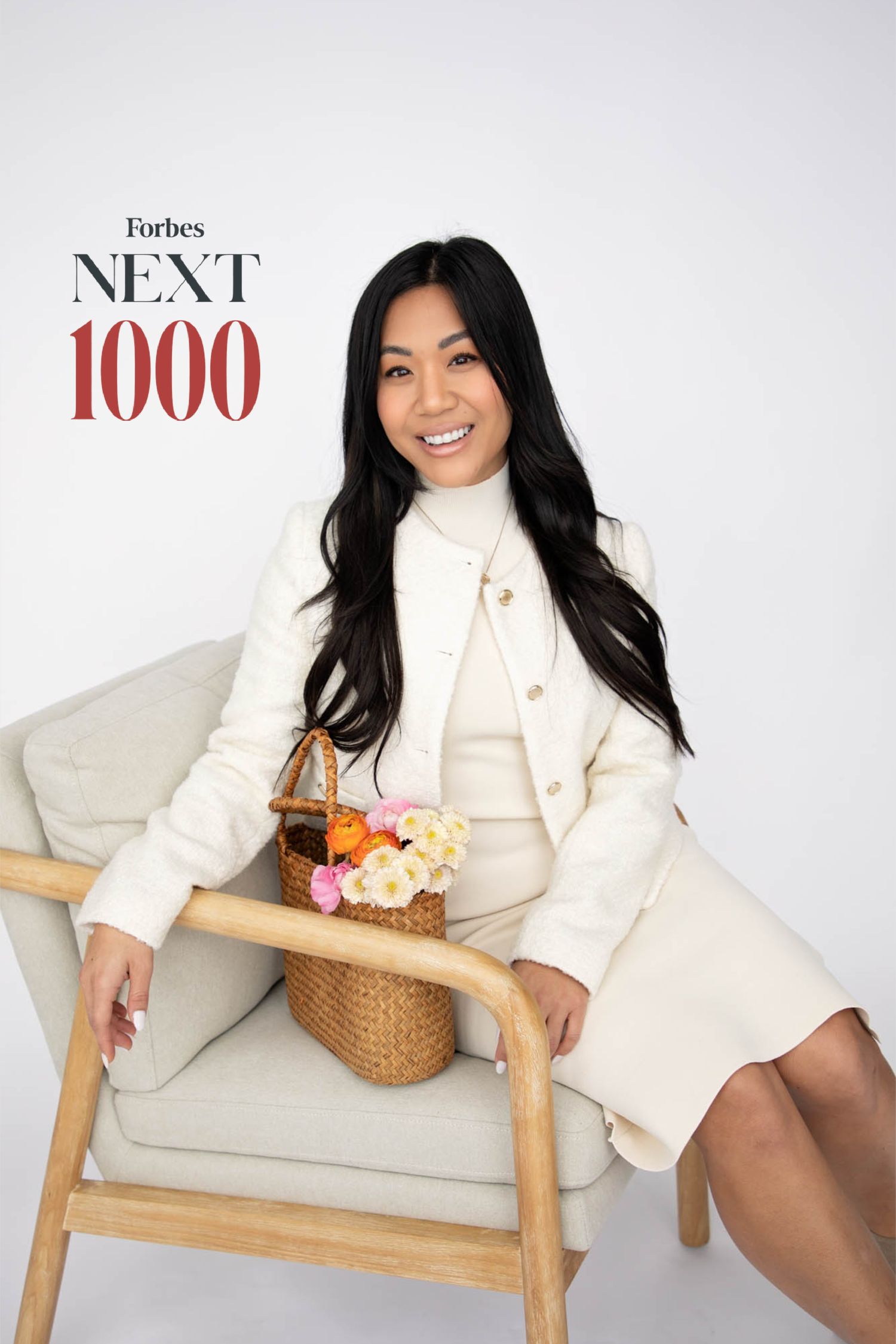 A woman sitting on a beige chair with a woven basket of flowers, smiling, against a white background with "Forbes NEXT 1000" text.