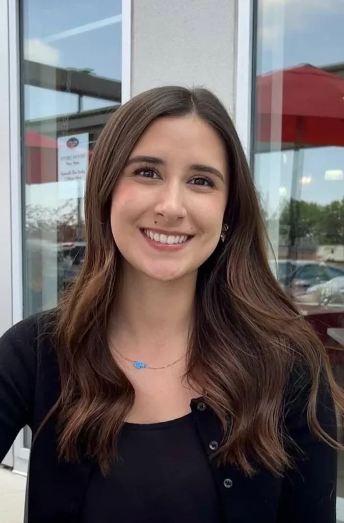 A young woman with long brown hair and a black shirt, smiling at the camera, outdoors in front of a glass building with a red umbrella and parking lot in the background.
