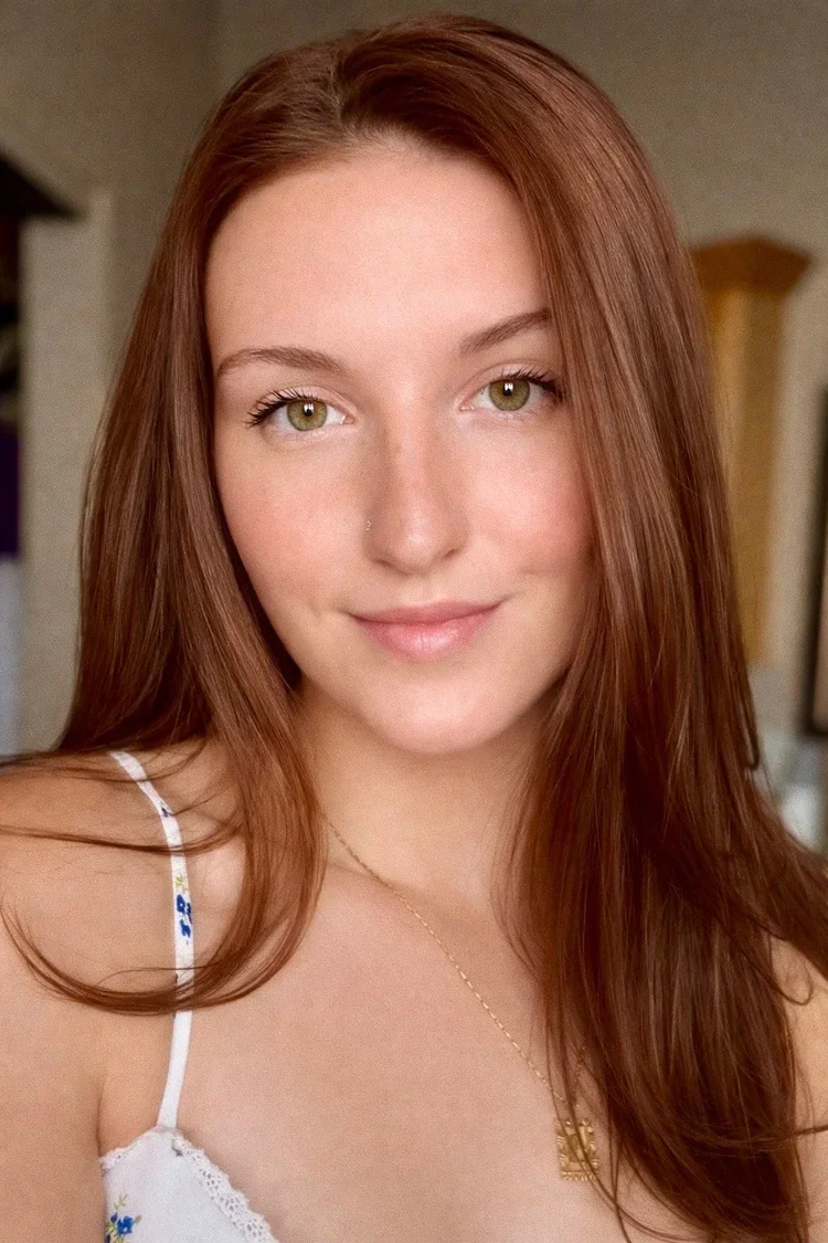 Close-up portrait of a young woman with long red hair, light skin, and green eyes, smiling gently at the camera.