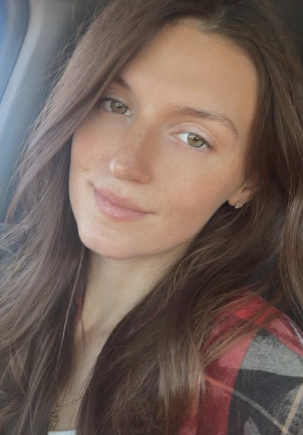 A woman with long brown hair and light-colored eyes smiling slightly, wearing a red and black plaid shirt, sitting in a vehicle.