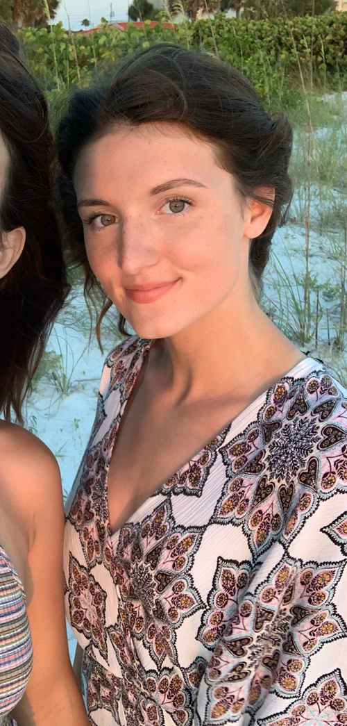 A young woman with light skin, green eyes, and short dark hair, smiling, standing outdoors near water and greenery.