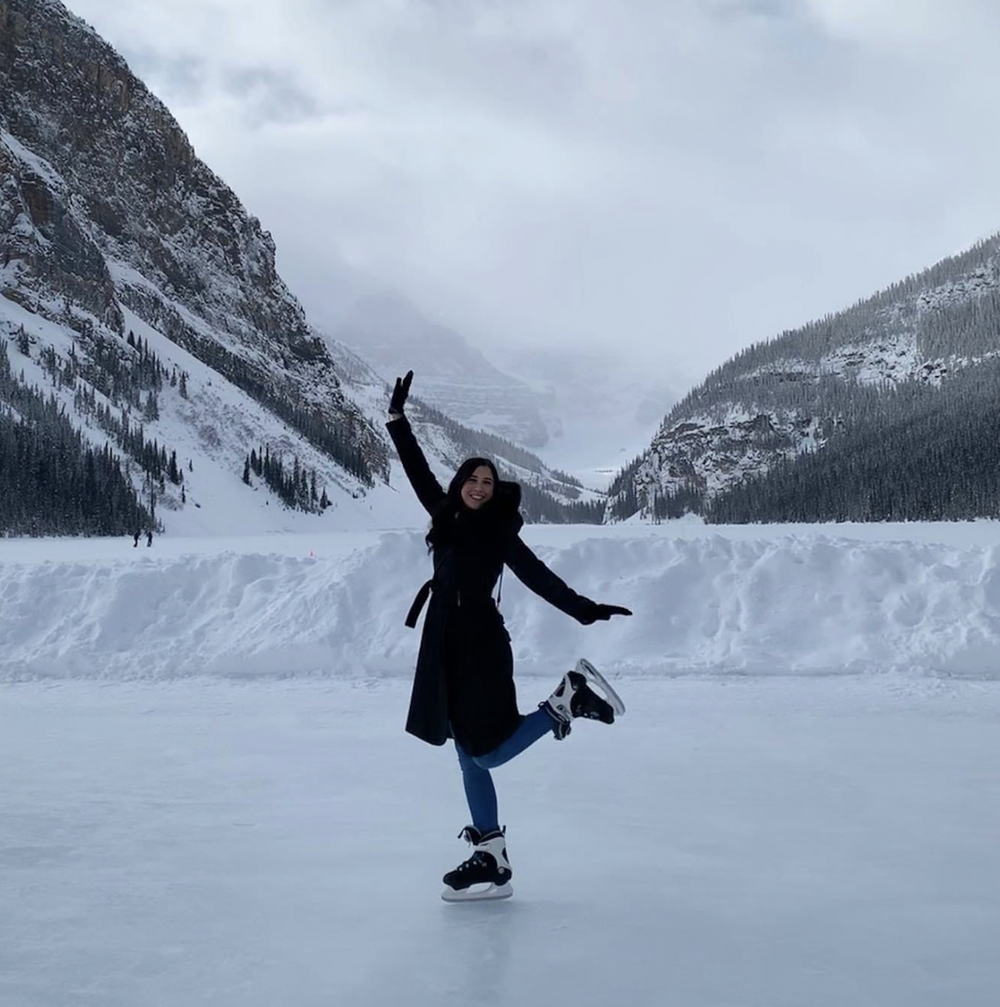 A woman ice skating on a frozen lake in a snowy mountain landscape with pine trees and cloudy sky.