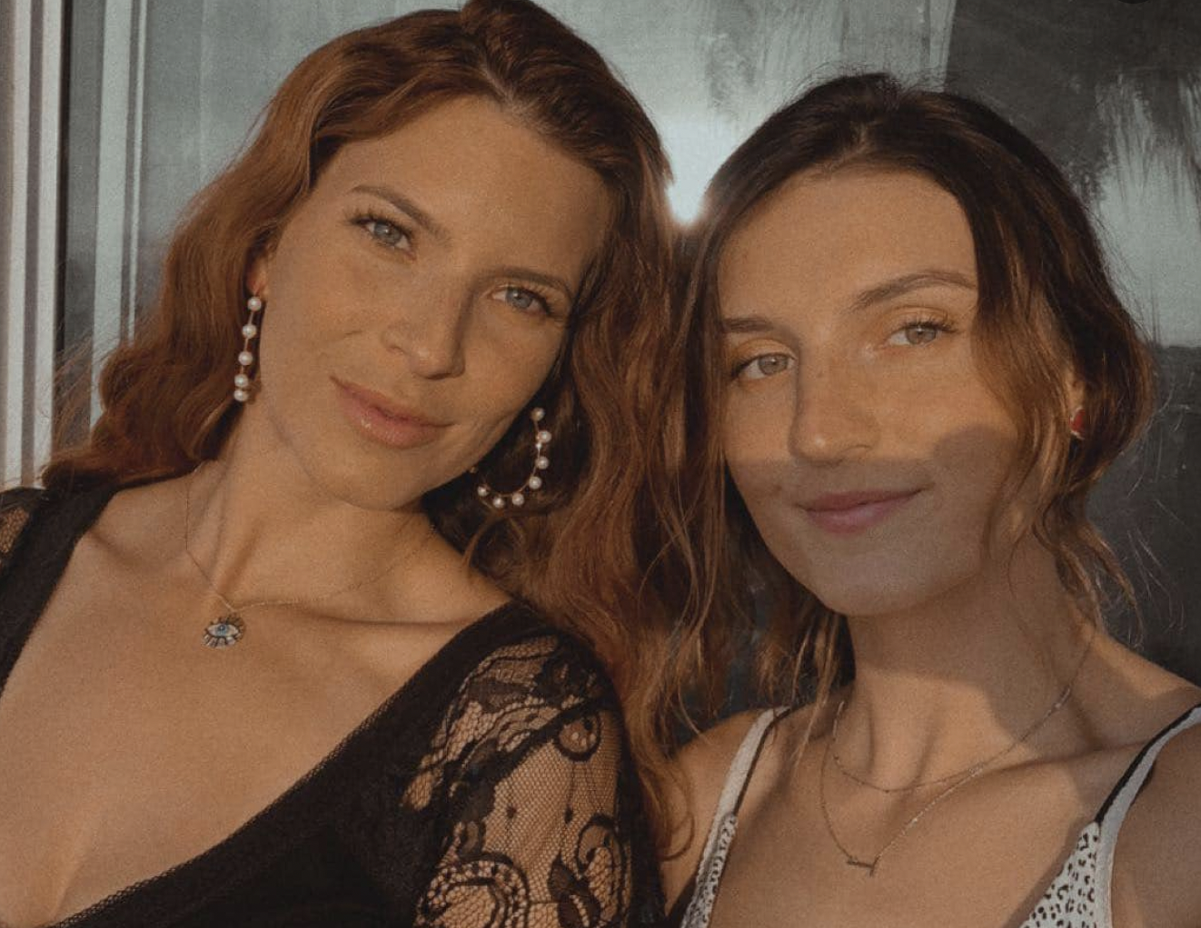 Two women taking a selfie, both smiling at the camera, with natural makeup and wavy hair. The woman on the left has red hair, hoop earrings, a necklace with an eye-shaped pendant, and is wearing a black lace top. The woman on the right has brown hair