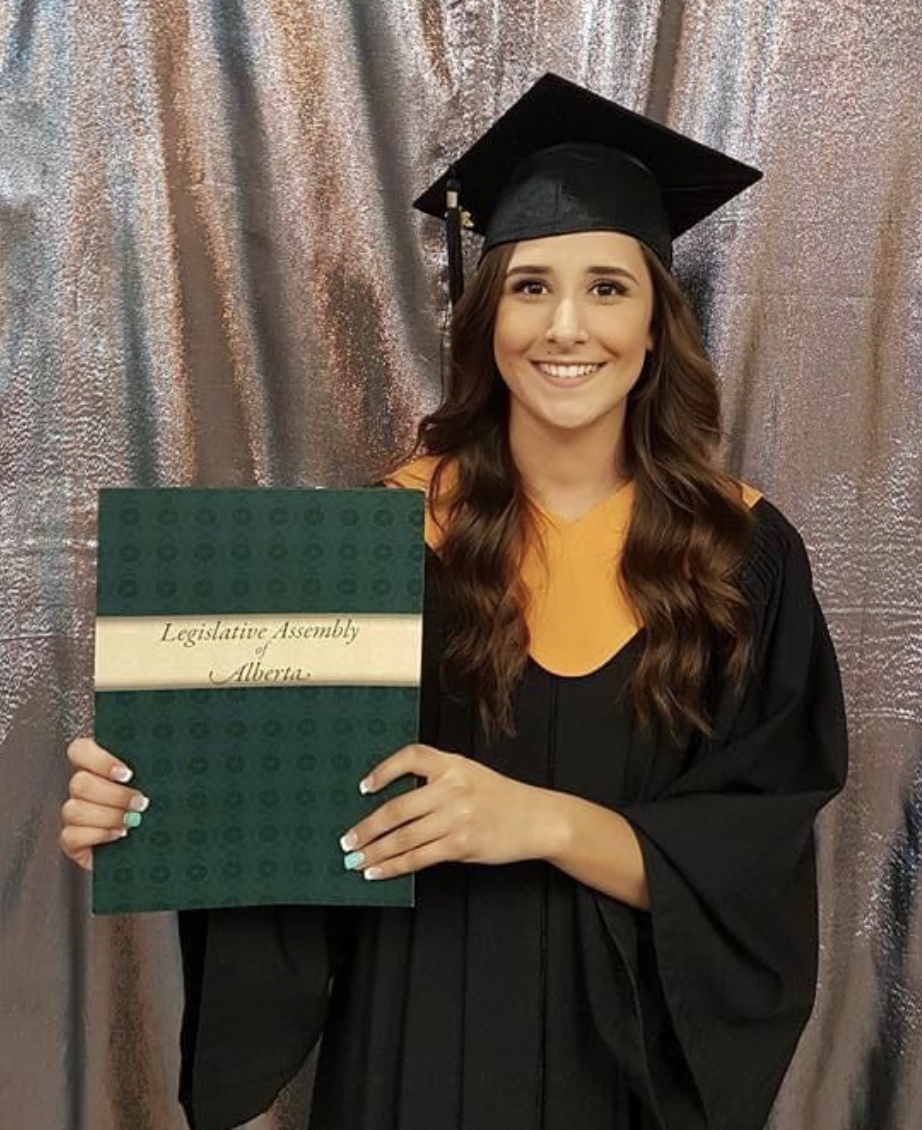 A young woman in a black graduation gown and cap holding a diploma cover that says "Legislative Assembly of Alberta," standing in front of a bronze-colored, textured backdrop.