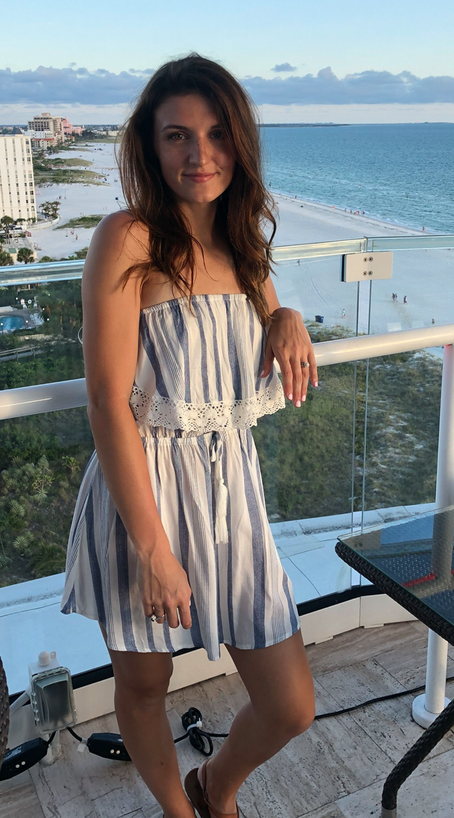A woman with long brown hair in a strapless striped dress standing on a balcony overlooking a beach and ocean.