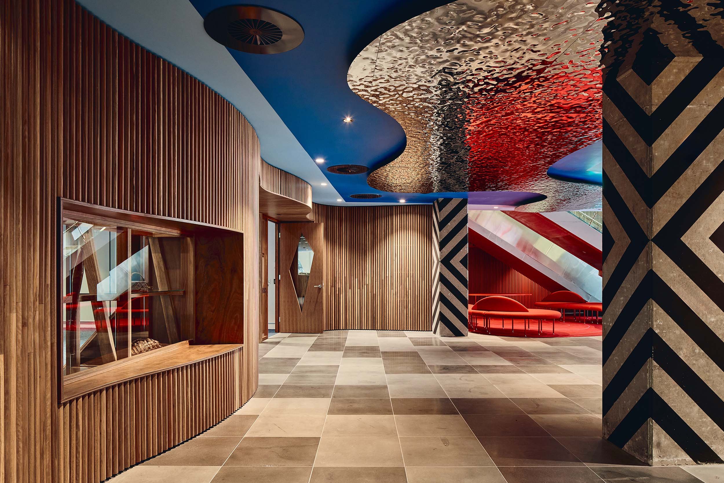 Texture and colour are on show in this foyer space of KHT. Warm timber wall panelling sit against a bright blue ceiling that features textural mirrored panels that evoke the nearby Yarra River. Patterned concrete pillars and bold red carpet and seats