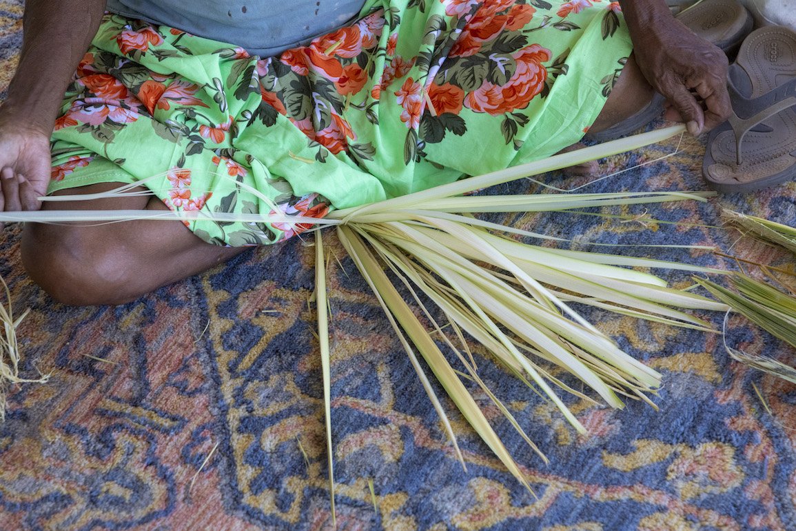 Merrepen (sand-palm) being hand woven by someone wearing a bright floral printed skirt, sitting with their legs crossed on a patterned rug.