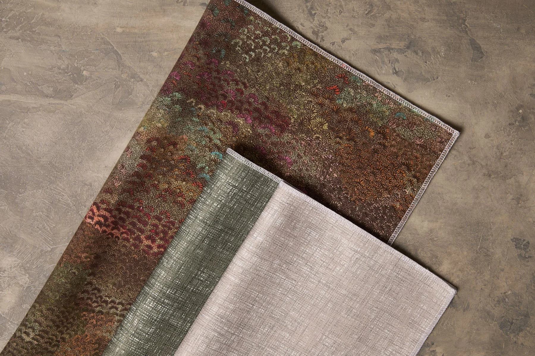 Three patterned fabrics laid out and folded on a flat surface, in earthy pink, green and taupe colours.