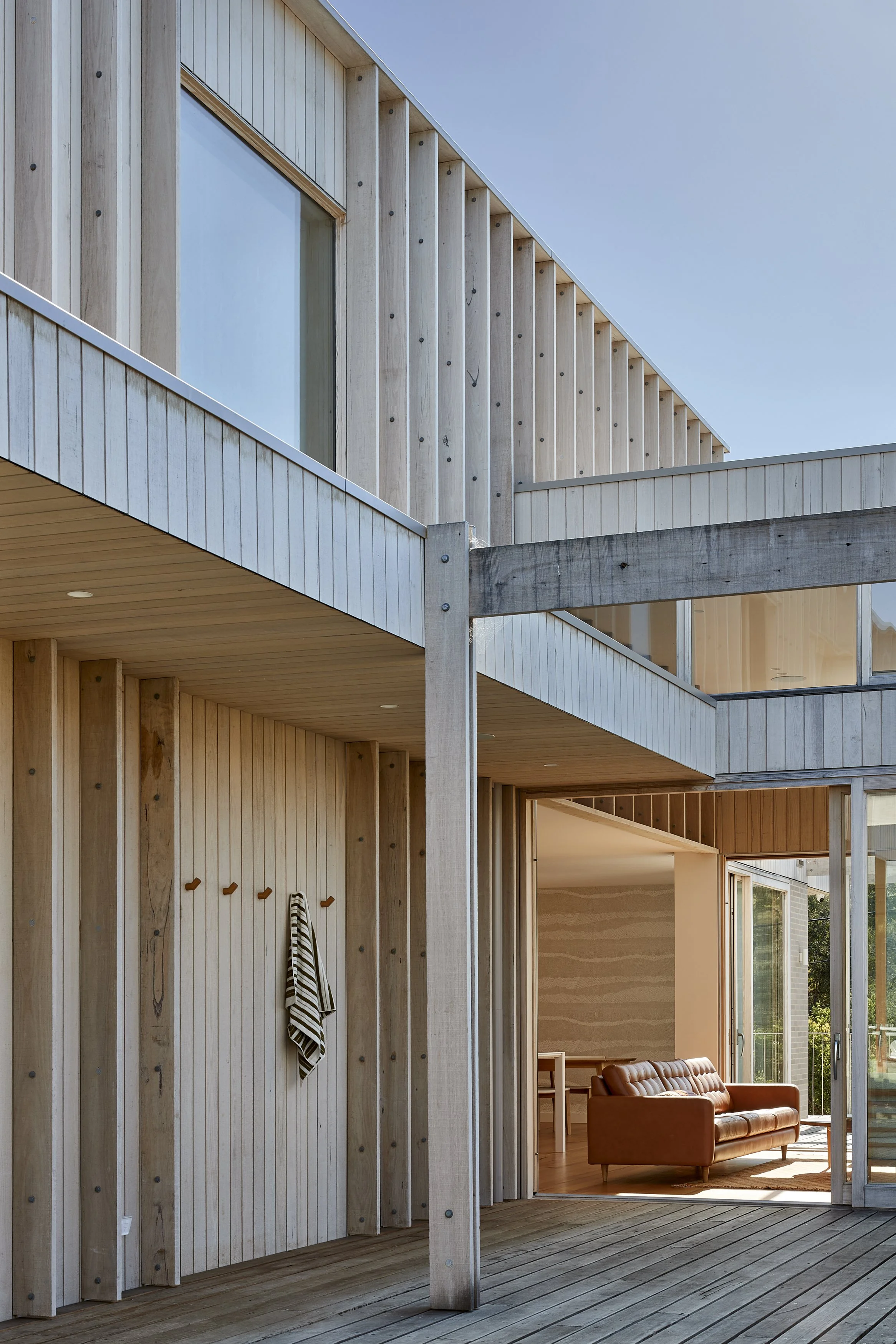 Blairgowrie Beach Club's exterior in pale Australian timber with large windows give a coastal feel. A large sliding door is open to the lounge room where sun is shining on the tan leather couch.