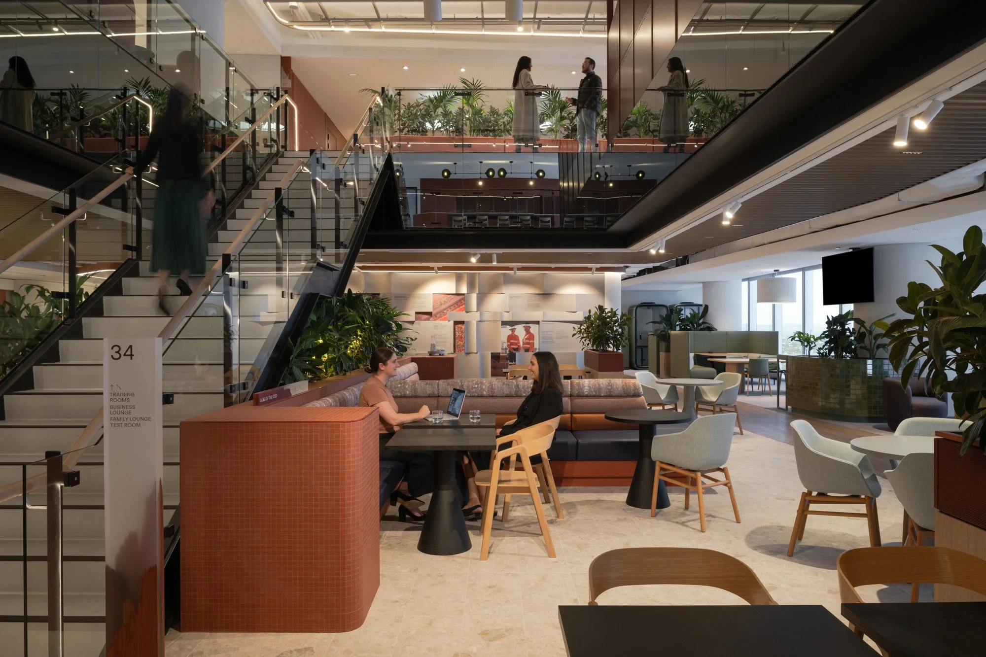 The foyer of BHP Perth offices. A vibrant workplace environment featuring a multi-level atrium design with prominent staircases, abundant greenery integrated into the seating areas, and a warm, textured colour palette.