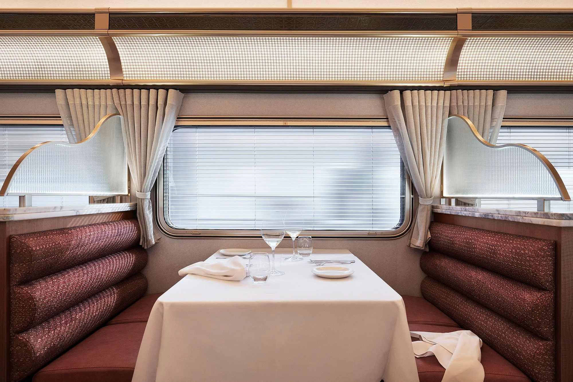 Dining booth with maroon upholstered benches, a white tablecloth, wine glasses, and place settings in front of a window with horizontal blinds and beige curtains, in a train dining car.