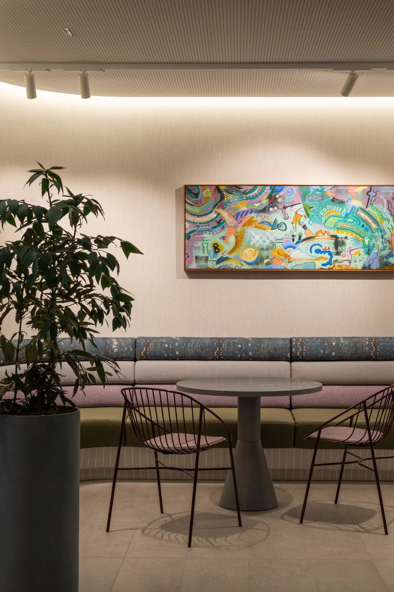 A collaborative workspace within BHP Perth Office. Printed Banquette seating in tones of dark blue, grey, lilac and green, sitting with charcoal tables, chairs and indoor planters. A colourful abstract painting sits above the banquette seating.