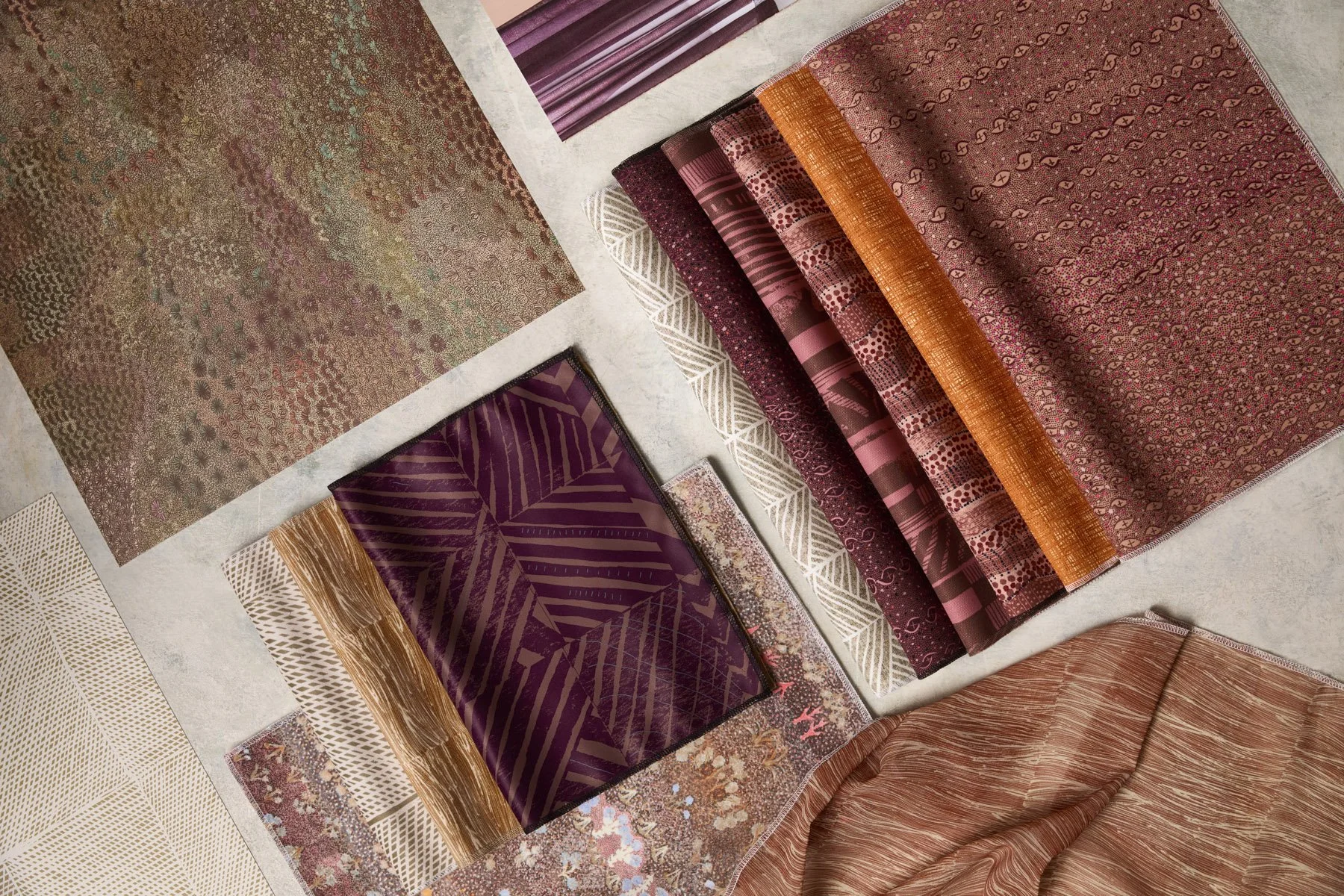 Various patterned fabrics and wallpapers laid out and folded on a flat surface, in earthy pink, plum and brown colours.