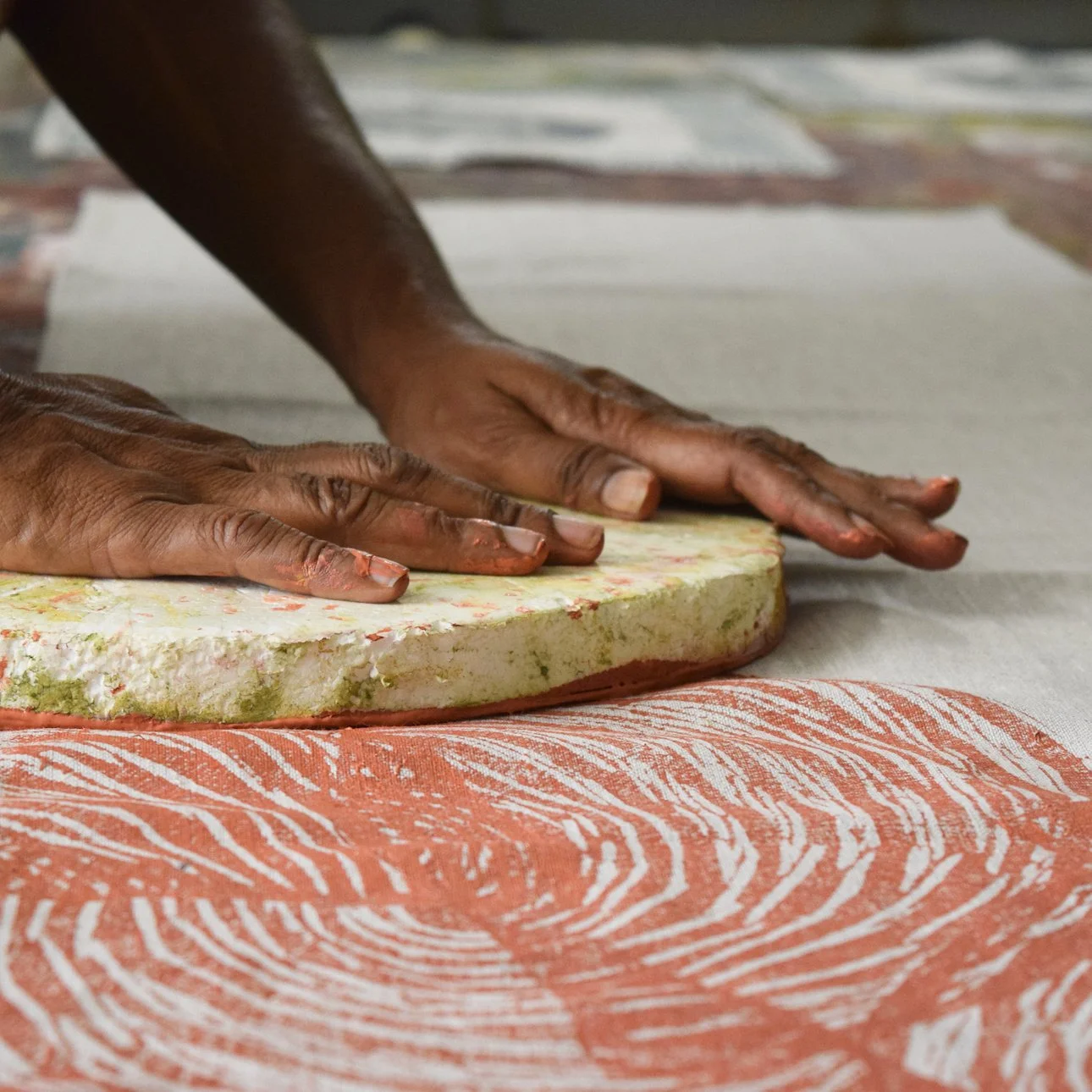 Interior art studio with close up of two hands of First Nations woman pressing a wood block print onto white fabric, print is terracotta colour.