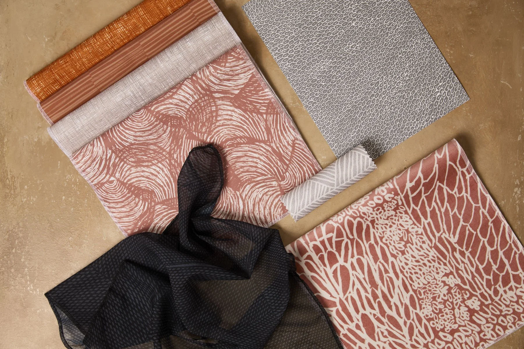 Various patterned fabrics and wallpapers laid out and folded on a flat surface, in terracotta, grey and black colours.