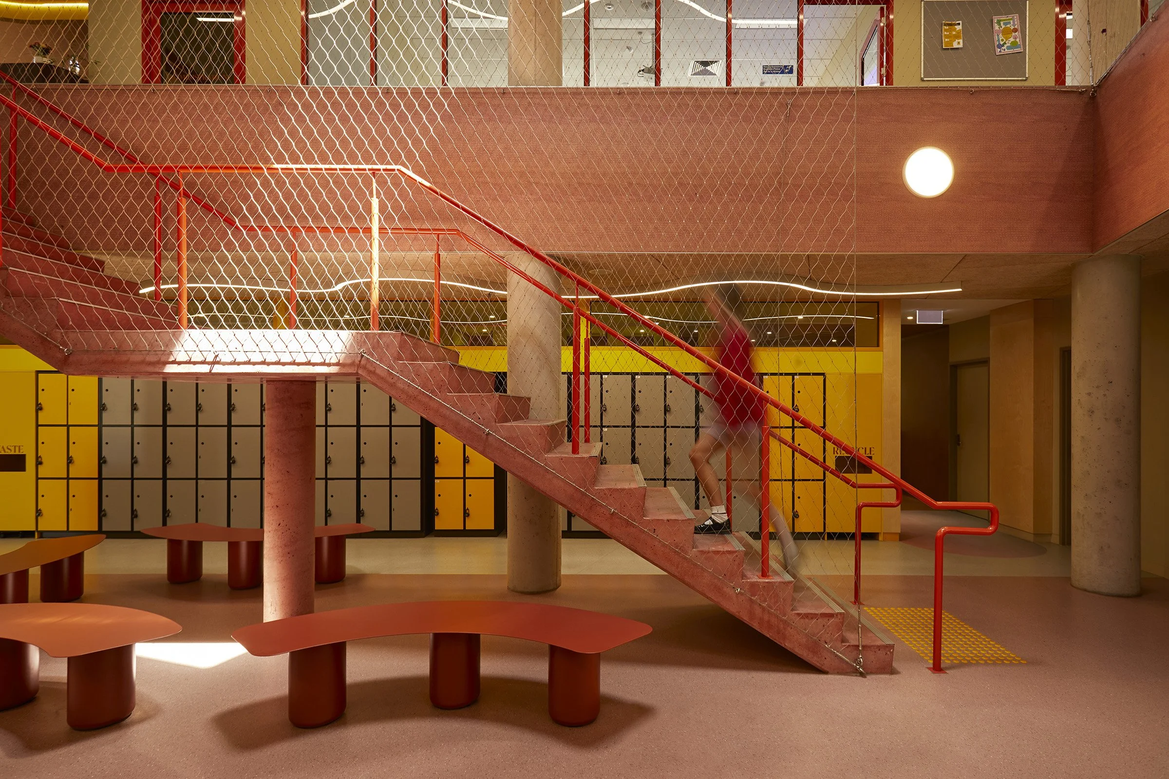 Interior of a school or recreational facility with a staircase enclosed by a security net. Yellow lockers line the back wall, and there are round tables in the foreground. A blurred figure descends the stairs.