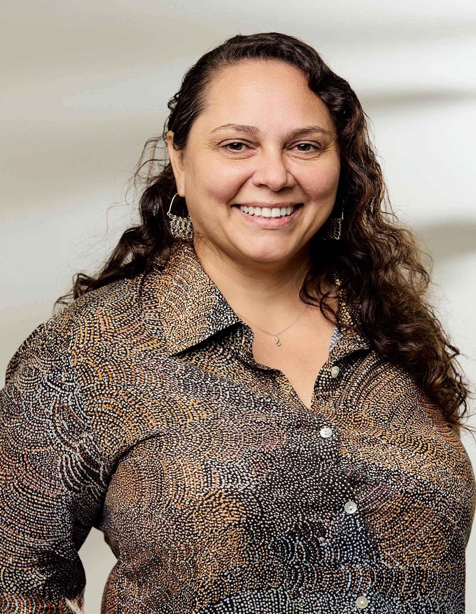 A woman with long curly brown hair, wearing a brown, black, and white patterned button-up shirt, smiling and standing against a plain light background.