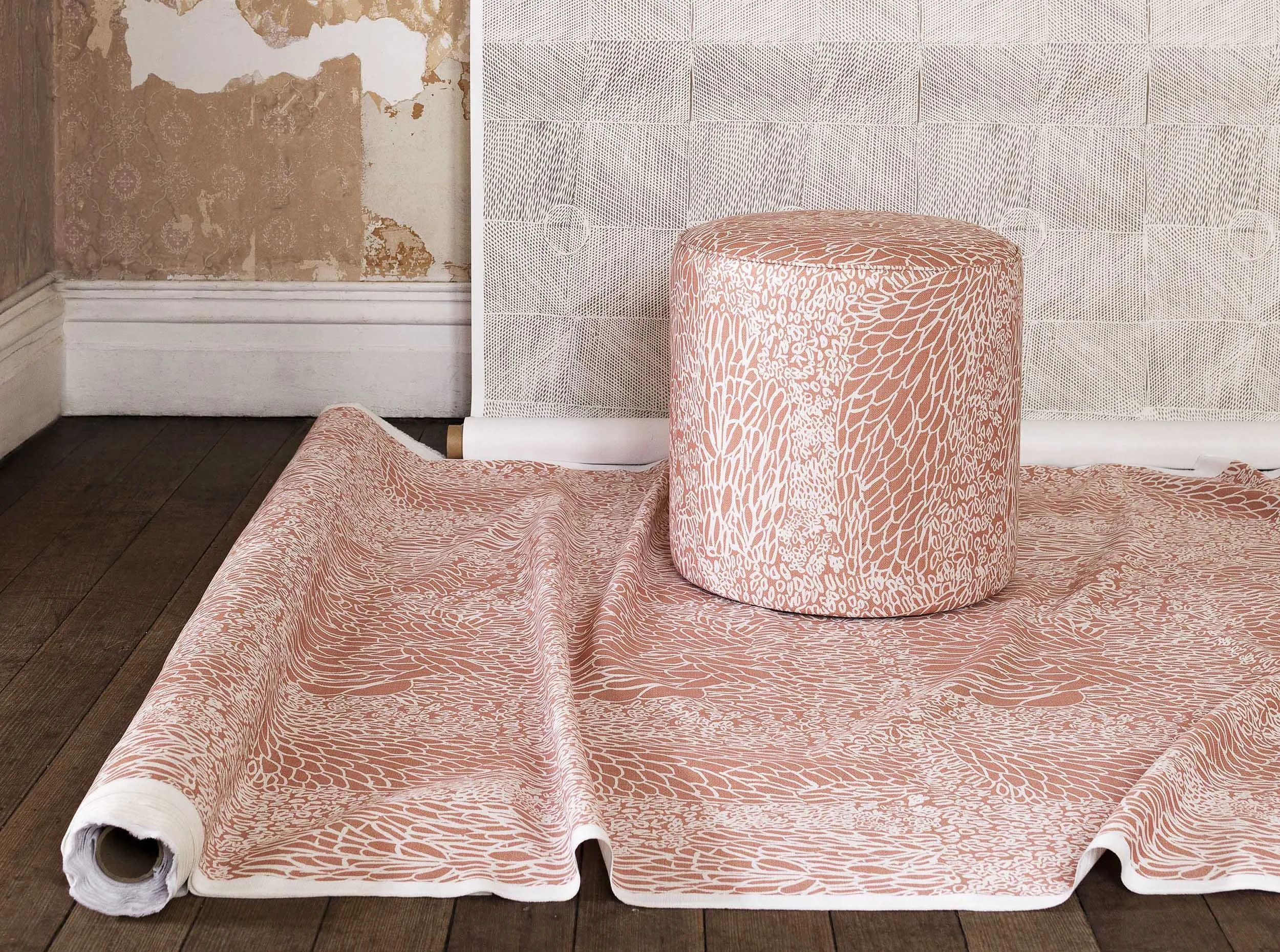Interior image of round ottoman sitting on fabric laid out on floor in foreground, background has wallpaper hanging from top, patterns featuring First Nations designs colours include terracotta, white and green