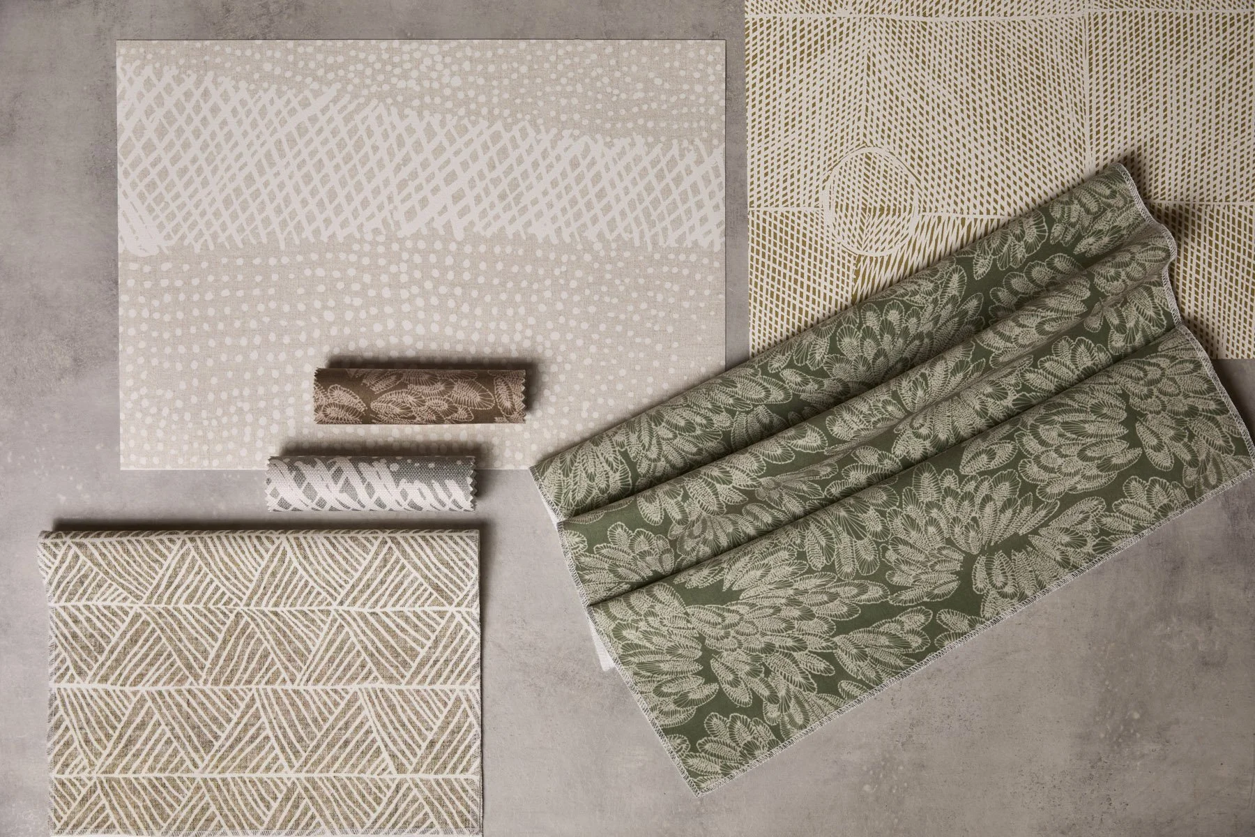 Various patterned fabrics and wallpapers laid out and folded on a flat surface, in grey, green and brown colours.