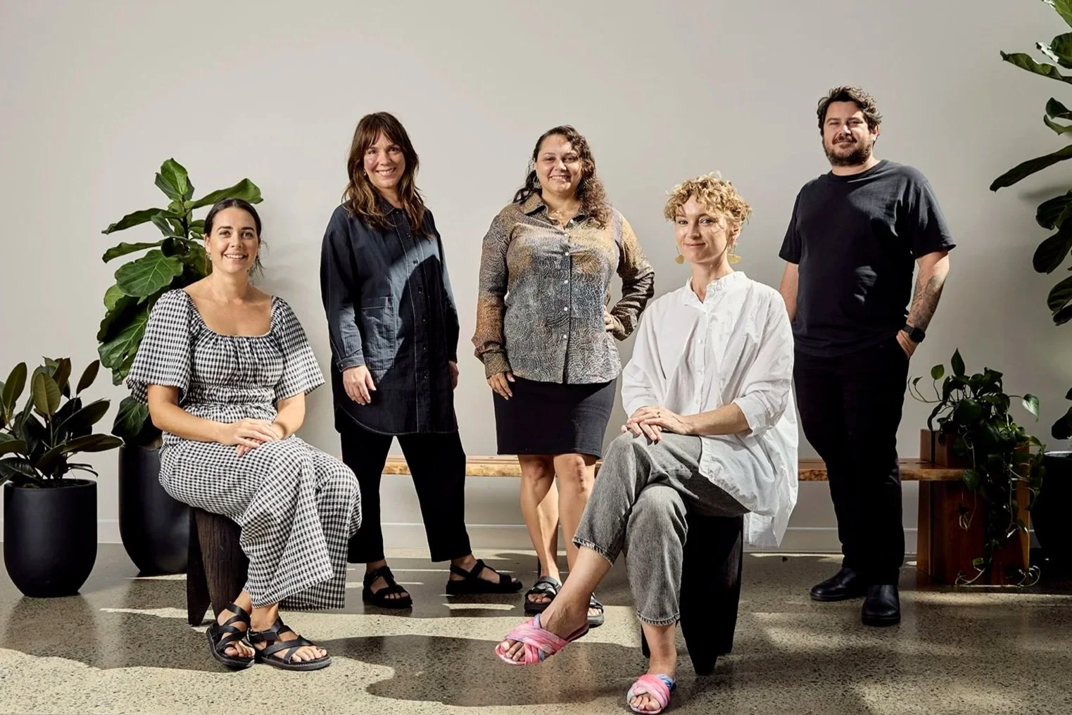 Group of five diverse adults, four women and one man, posing together indoors with large potted plants and a white wall background.