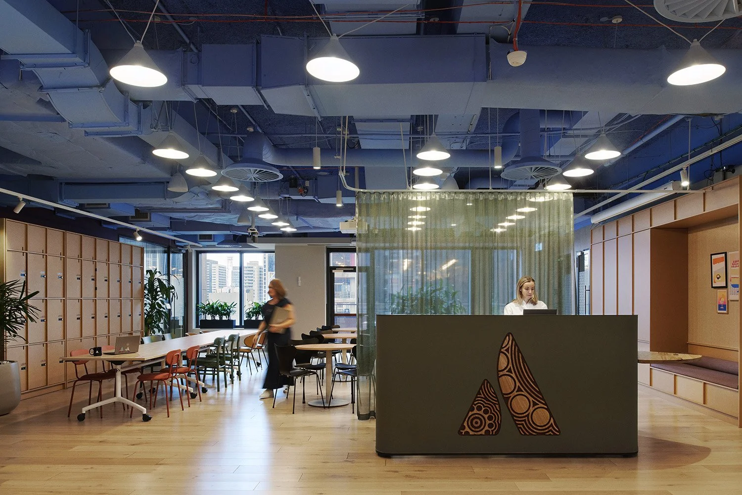 The open plan reception space of Atlassian's office. Timber flooring, a green reception desk featuring a decorative 'A',, semi-sheer green printed curtain divider, industrial style overhead pendant lights and blue exposed services on the celling.