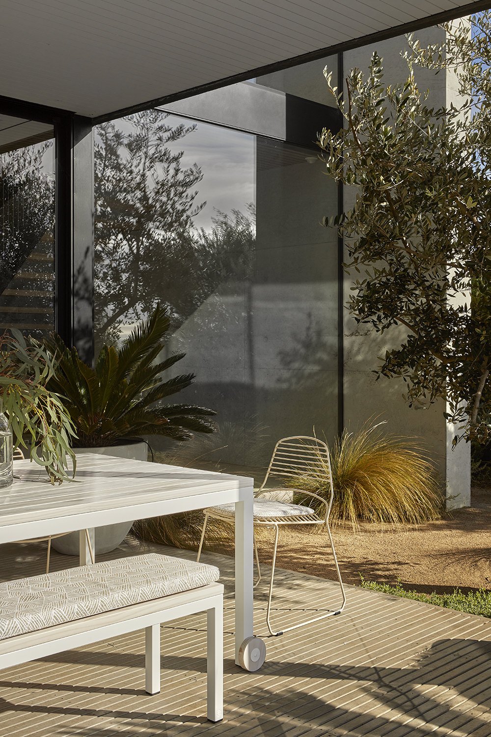 Modern outdoor patio with white table and bench, metal chair, potted plants, and trees reflected on glass wall.