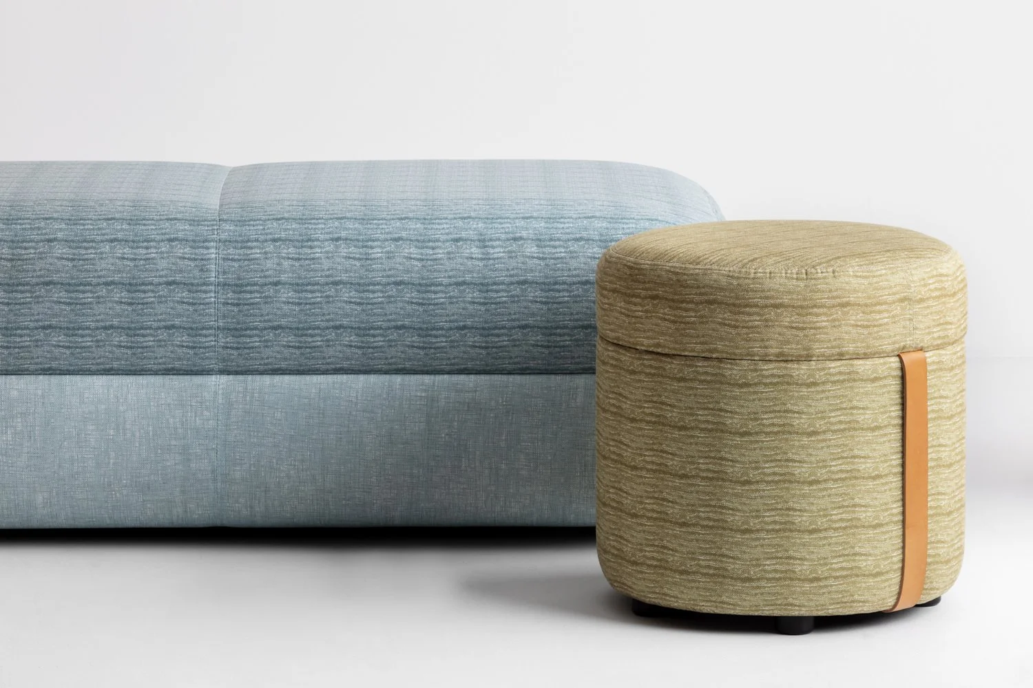 A light blue upholstered sofa next to a beige fabric ottoman with a leather strap decoration, on a white background.