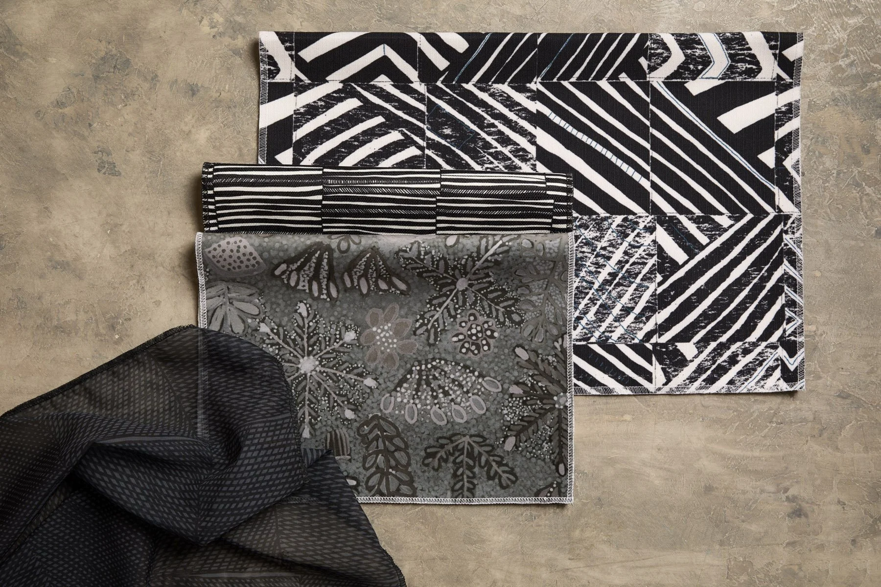 Various patterned fabrics laid out and folded on a flat grey surface, in moody black, green and white colours.