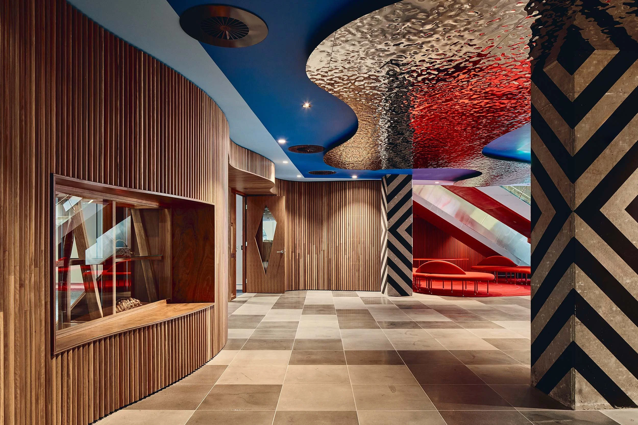 Texture and colour are on show in this foyer space of KHT. Warm timber wall panelling sit against a bright blue ceiling that features textural mirrored panels that evoke the nearby Yarra River. Patterned concrete pillars and bold red carpet and seats