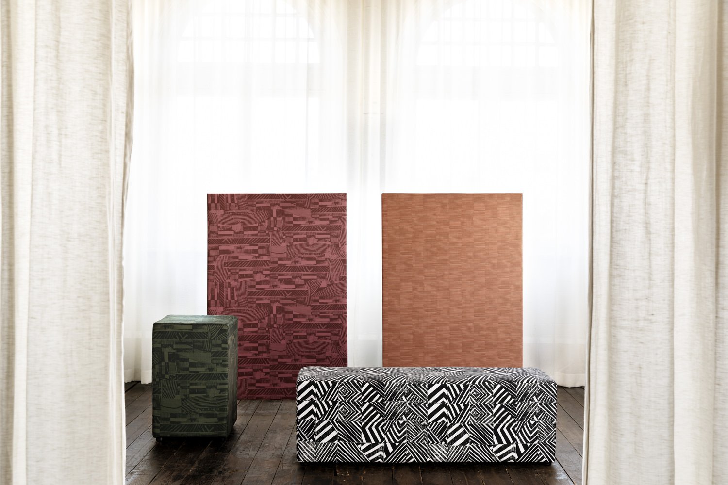 Interior view with two square panels at rear, rectangle ottoman in foreground, square ottoman at left, all upholstered with various patterns featuring First Nations designs. White sheer curtain in background and wooden floorboards.