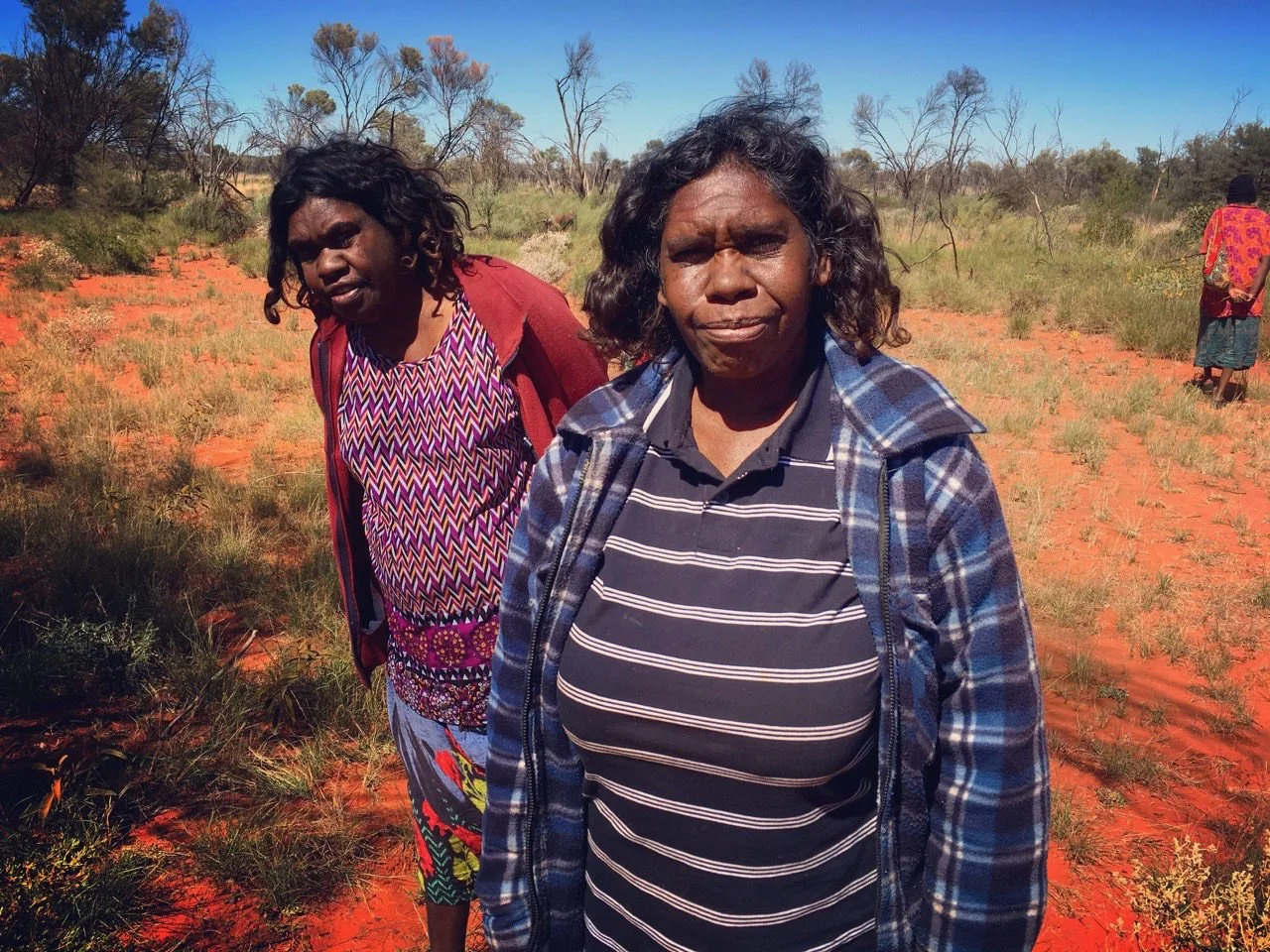 Two Indigenous female artists stand in the bush on red soil surrounded by trees, grasses and blue sky.