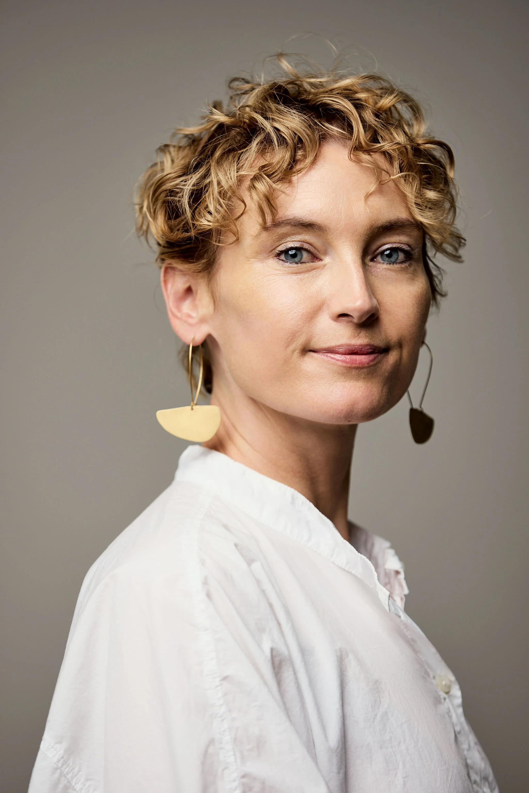 A woman with short, curly blonde hair and blue eyes, wearing large gold earrings and a white shirt, is looking at the camera against a neutral grey background.