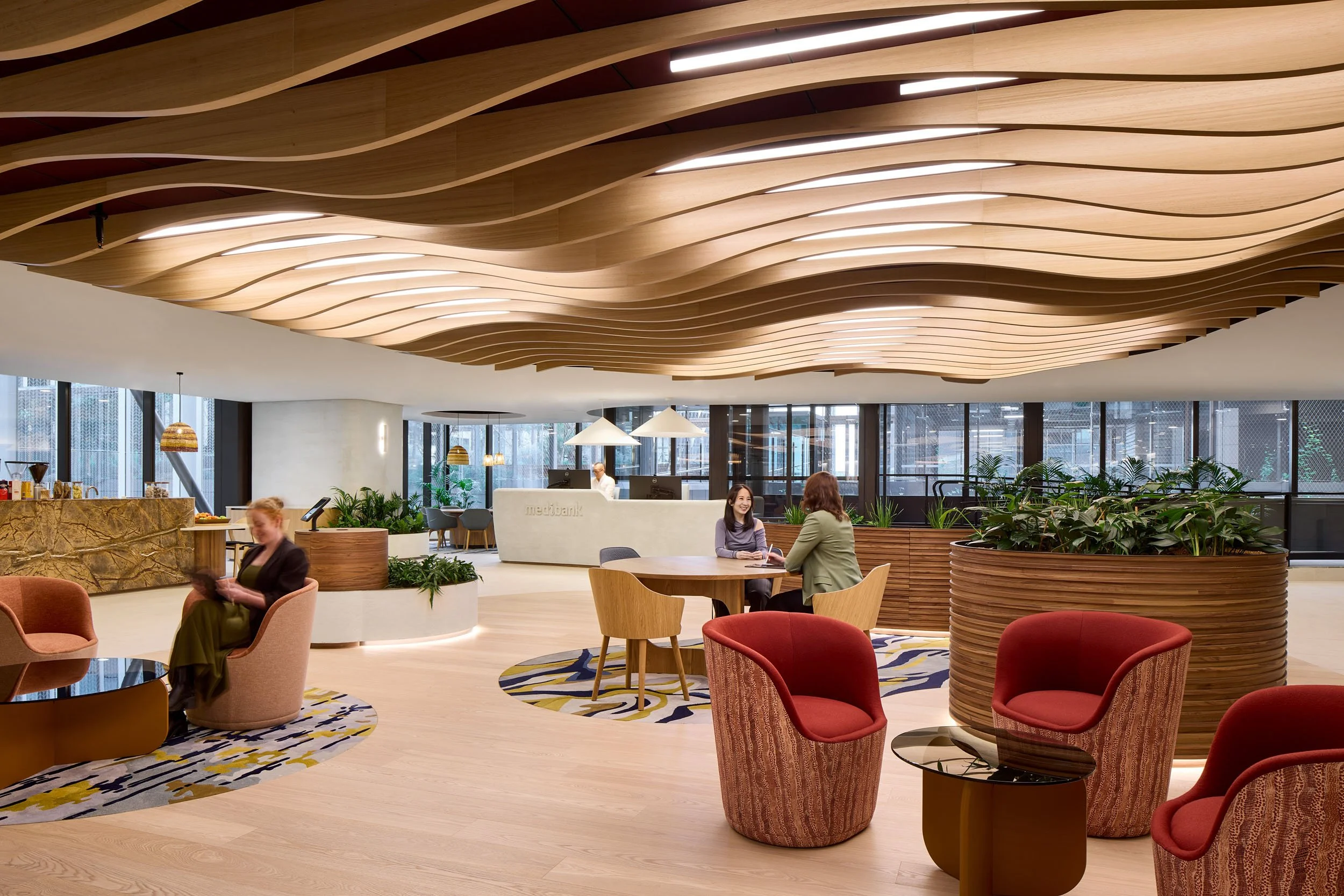 Modern office space for Medibank in Melbourne features a prominent custom wavy timber ceiling, large timber planters, patterned carpet and and upholstered armchairs in patterns with tones of red, peach and browns.