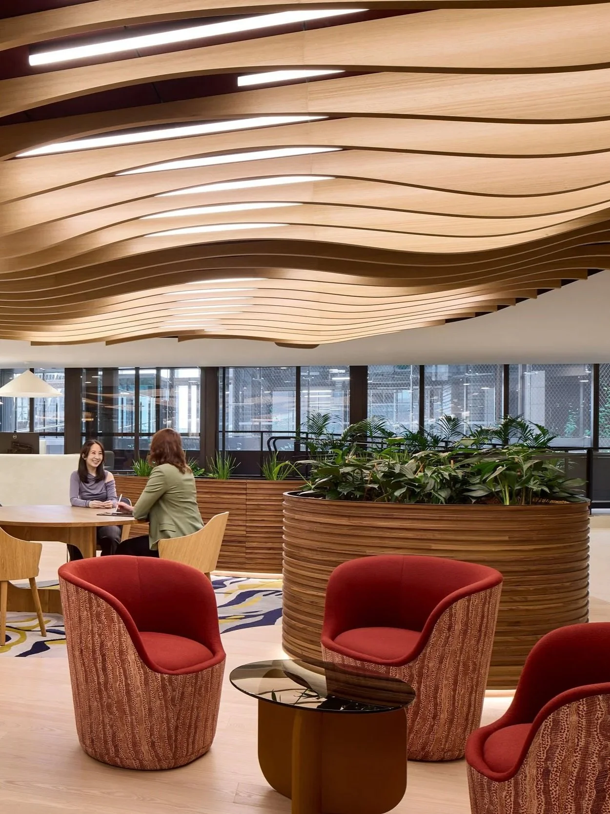 Modern office space for Medibank in Melbourne features a prominent custom wavy timber ceiling, large timber planters, patterned carpet and and upholstered armchairs in patterns with tones of red, peach and browns.