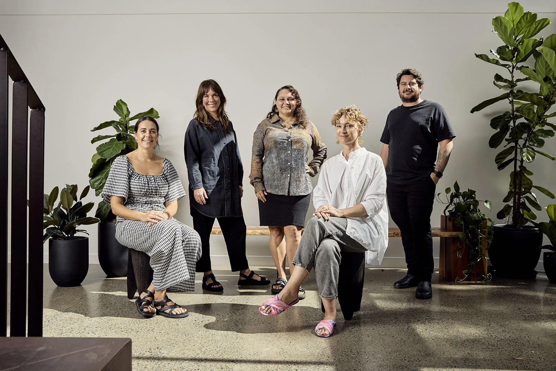 Group of five diverse adults, four women and one man, posing together indoors with large potted plants and a white wall background.
