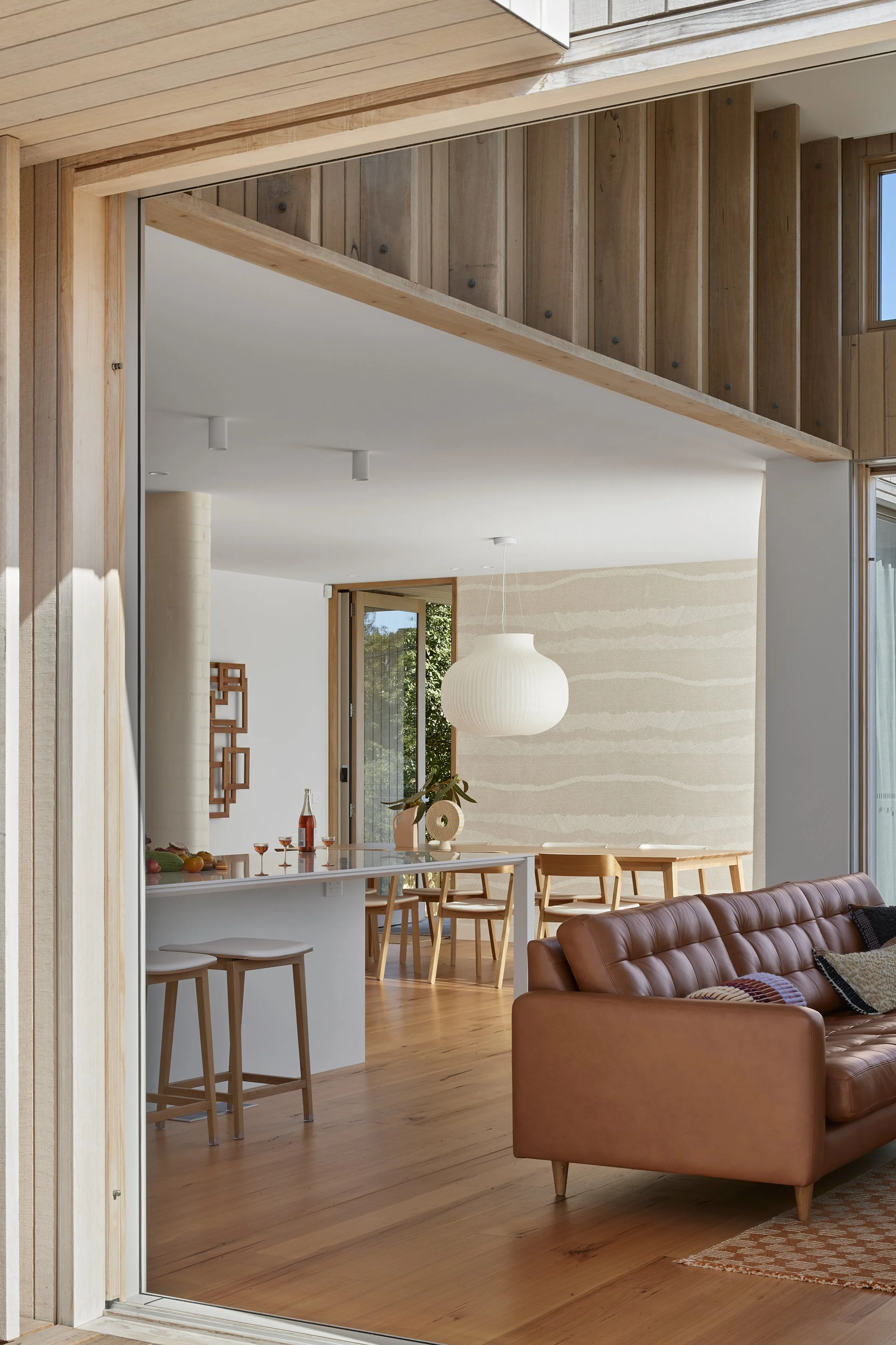 A view from the sliding door into the open plan kitchen/dining/lounge. A neutral colour palette includes timber, tan leather, cream upholstery and a stone coloured patterned wallpaper on the dining room wall.