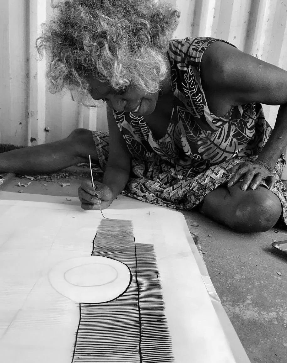 Black and white image of a First Nations woman sitting on ground painting an artwork, corrugated iron wall in background.