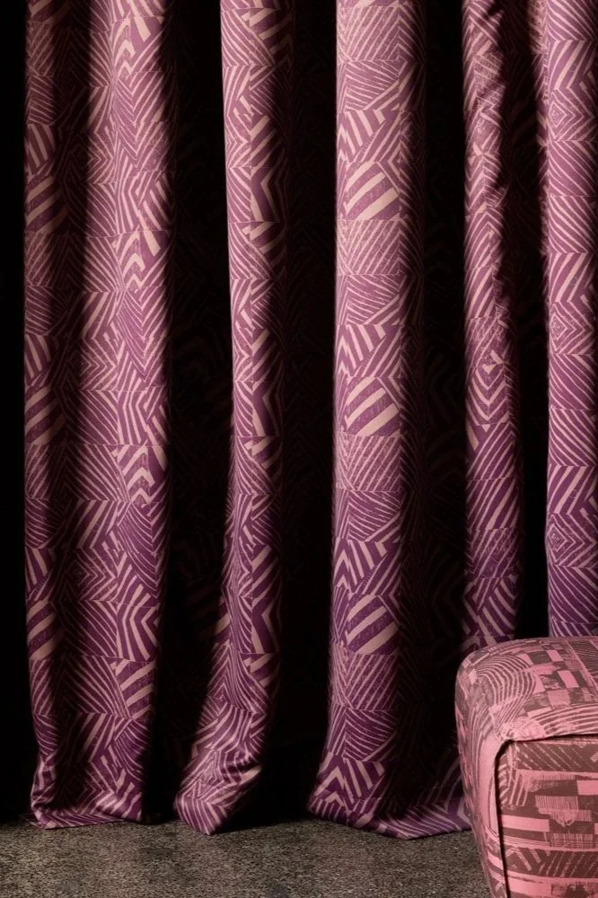 Purple curtains with a geometric white pattern hanging in front of a dark background, with a pink patterned armchair partially visible in the lower right corner.